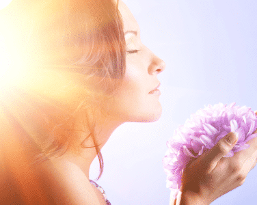 A woman holds a flower in front of her face, partially obscuring her features while standing against a blurred background.